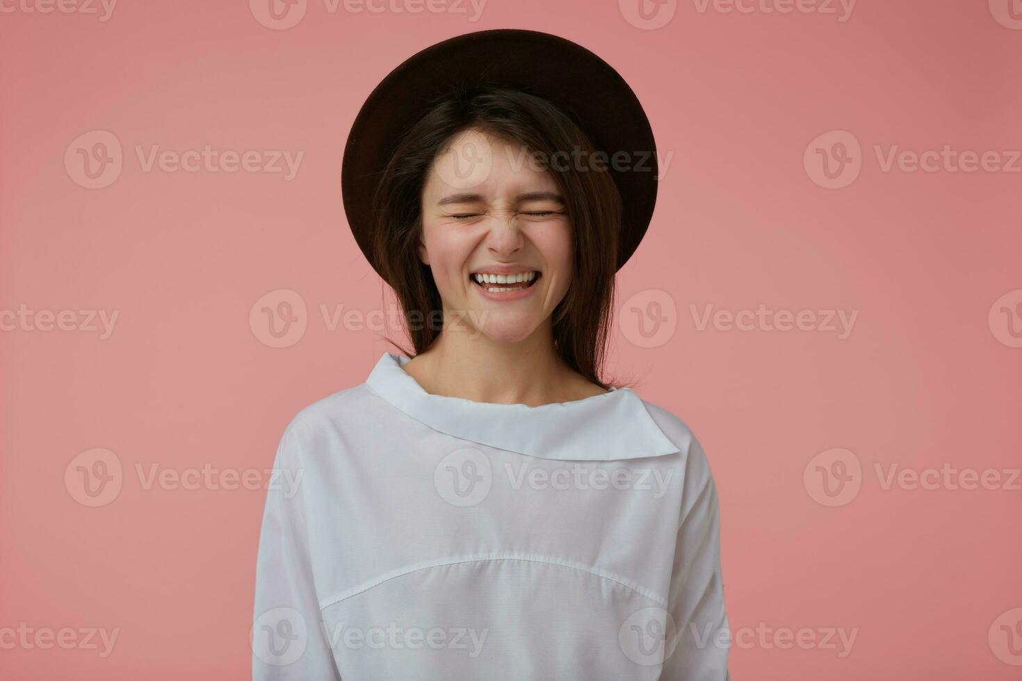 Teenage girl, happy looking woman with long brunette hair. Wearing white blouse and black hat. Laughing and keep eyes closed. Emotional concept. Stand isolated over pastel pink background photo