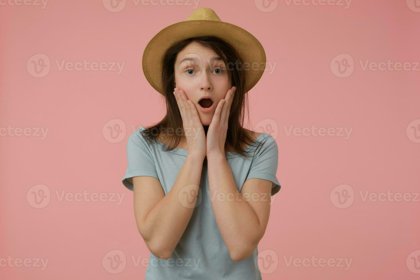 Adolescente chica, conmocionado mirando mujer con largo morena cabello. vistiendo azulado camiseta y sombrero. conmovedor su las mejillas, aterrorizado. acecho a el cámara aislado terminado pastel rosado antecedentes foto