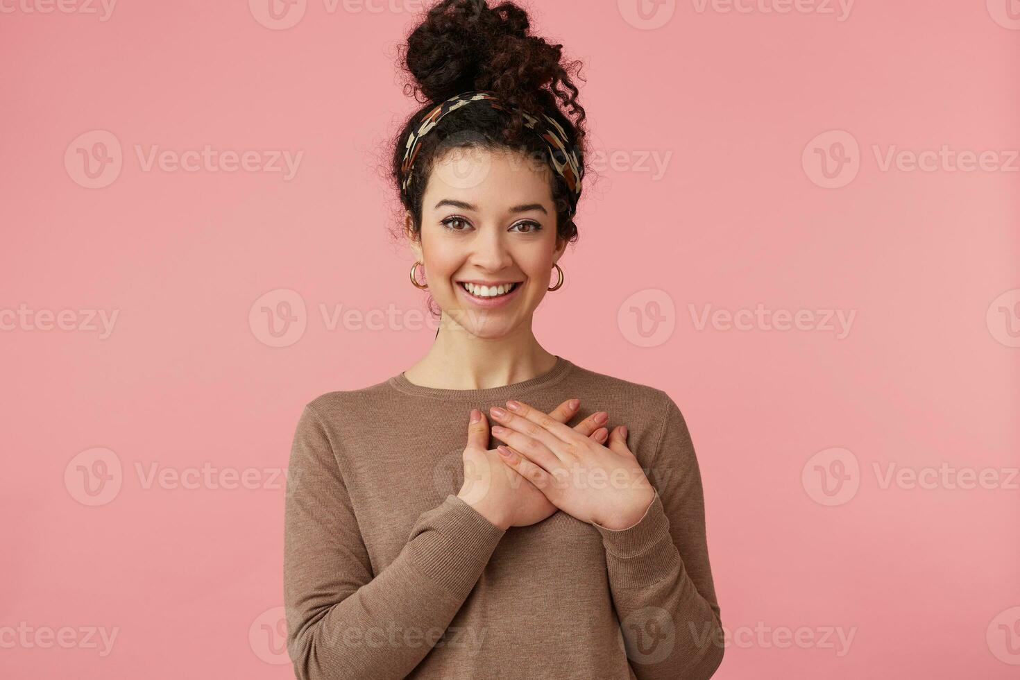 Portrait of a young beautiful curly girl, smiles broadly with hands to heart, hearing a complimentary, looking at camera isolated over pink background. photo