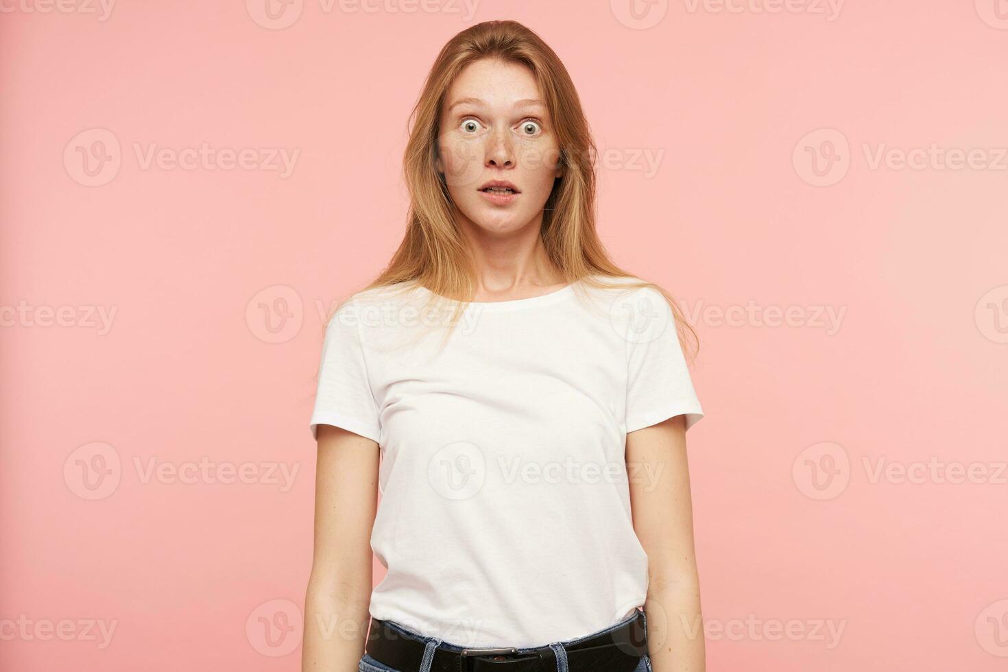 Studio photo of young amazed long haired redhead lady rounding surprisedly her green eyes while looking at camera, isolated over pink background in white basic t-shirt