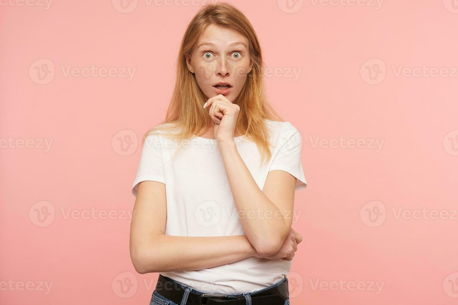 Studio photo of young bemused redhead female holding her chin with raised hand while looking surprisedly at camera with wide eyes opened, isolated over pink background