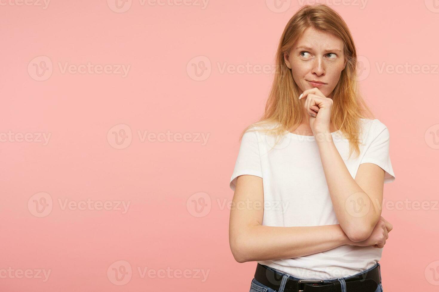 Puzzled young pretty long haired redhead lady holding her chin with raised hand and looking pensively aside, wearing casual clothes while posing over pink background photo