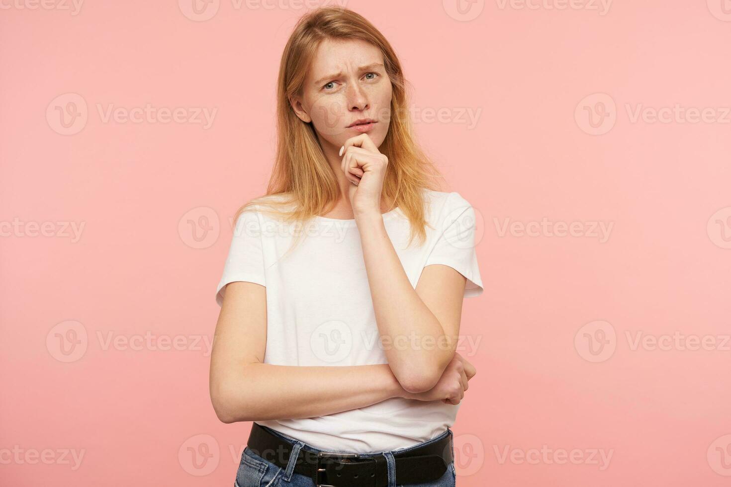 Pensive young attractive redhead woman with casual hairstyle keeping raised hand on her chin and squinting eyes while looking thoughtfully at camera, posing over pink background photo