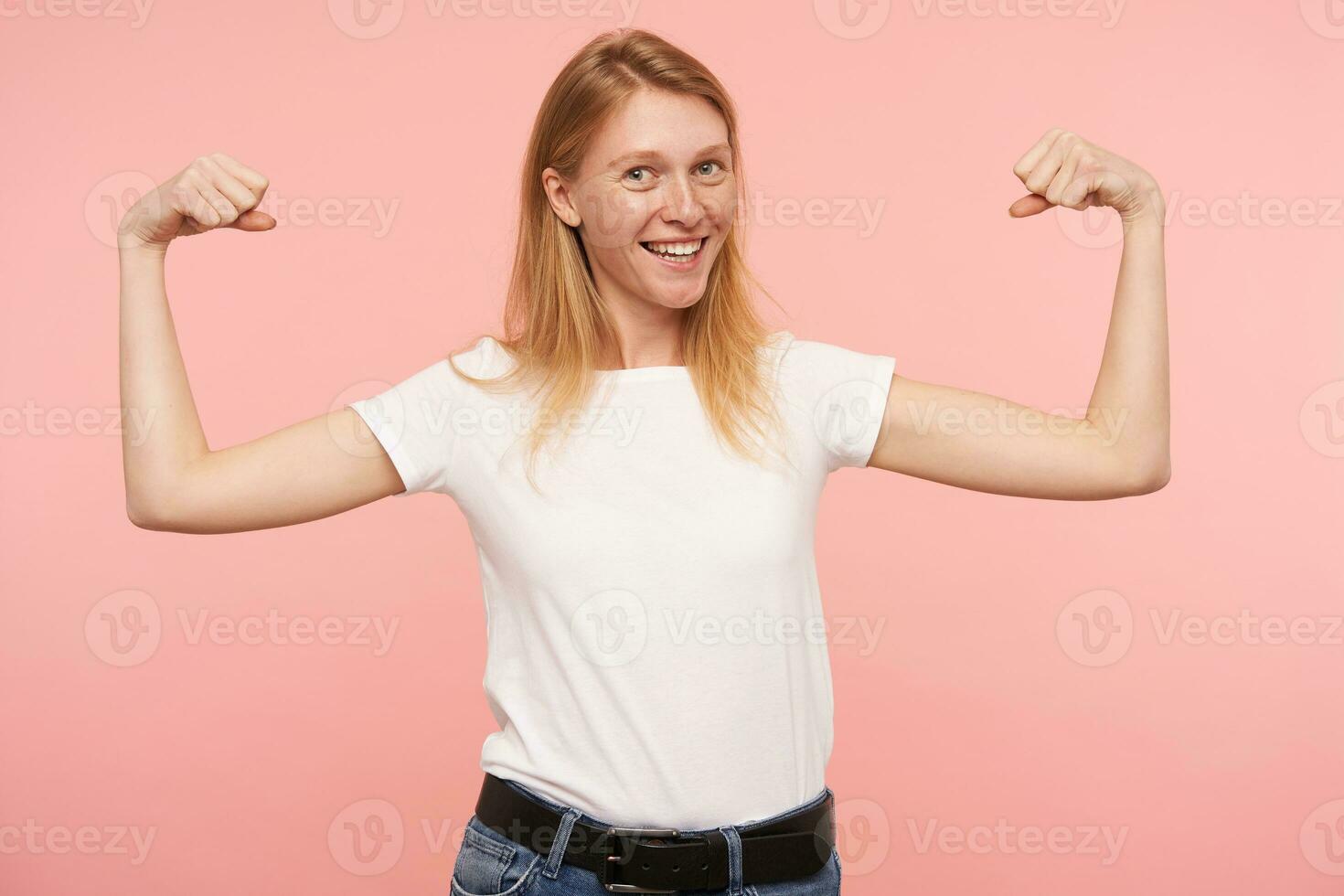 Indoor photo of young pretty redhead lady dressed in basic white t-shirt smiling broadly at camera while demonstrating her strong bicepses, posing over pink background