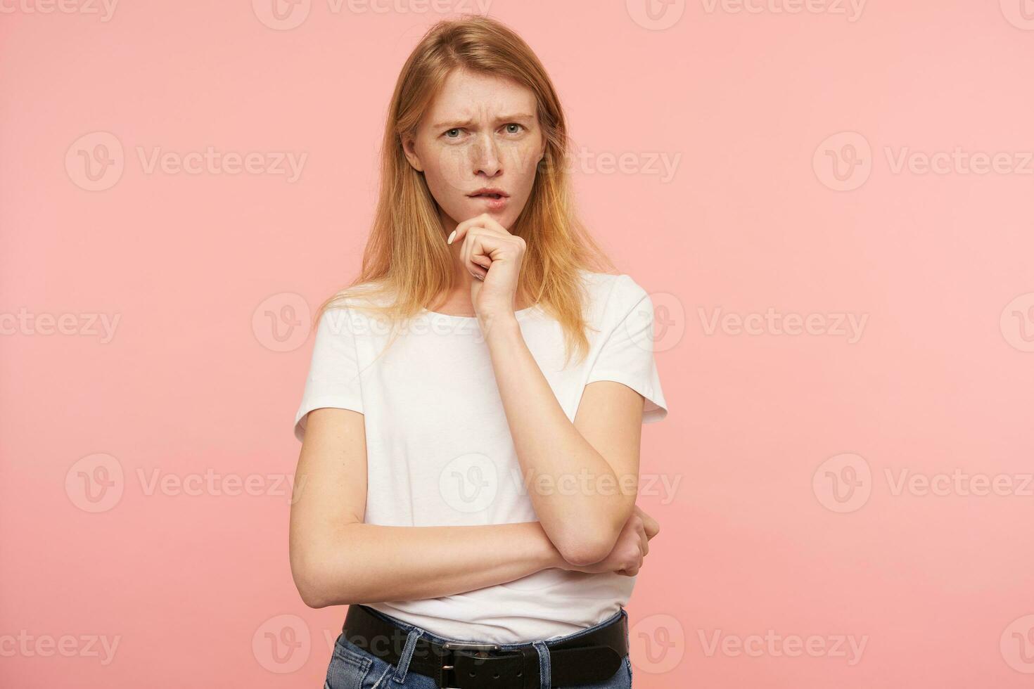 Serious young lovely green-eyed redhead female with natural makeup frowning her eyebrows while looking pensively at camera and keeping raised hand on her chin, isolated over pink background photo