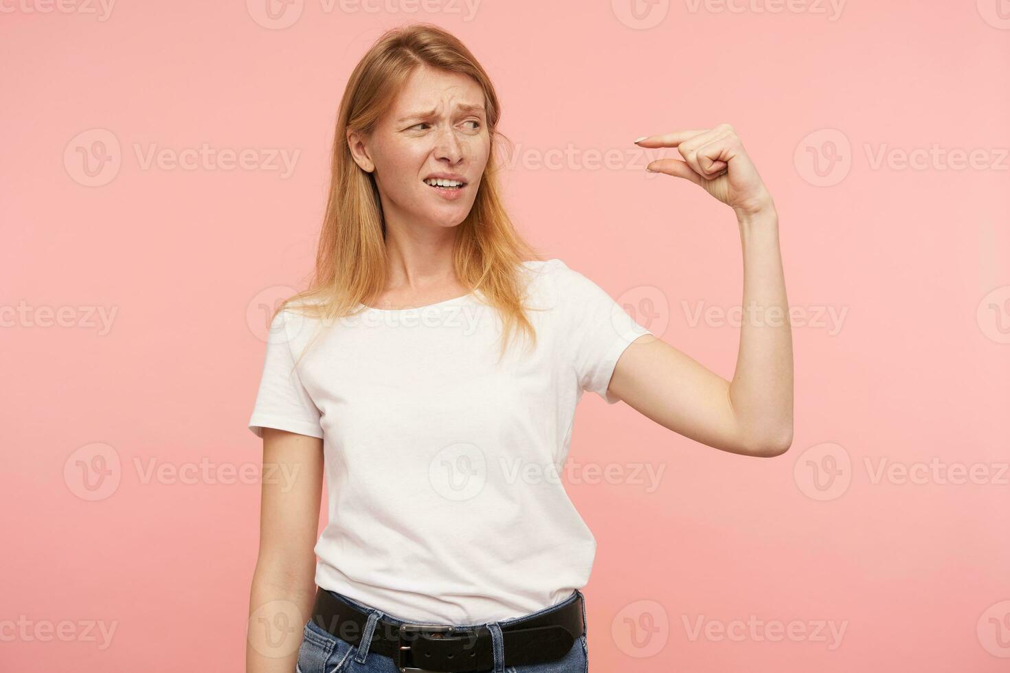 Disappointed young attractive long haired redhead woman grimacing her face while measuring something invisible with fingers, standing against pink background photo