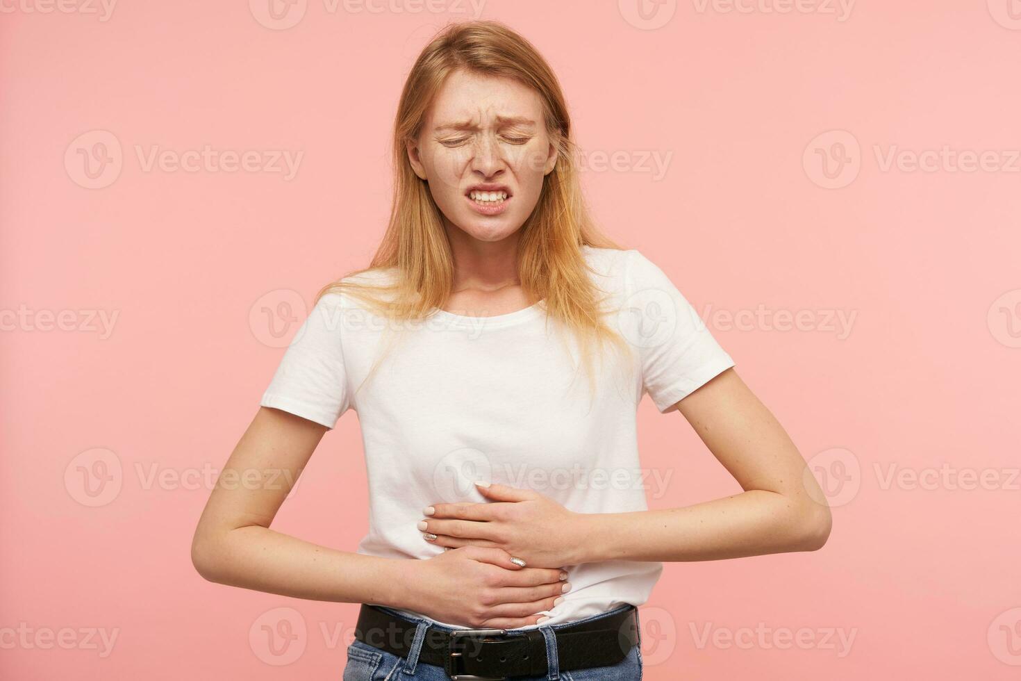 interior foto de joven bonito pelirrojo dama acuerdo su ojos cerrado y ceñudo cara mientras teniendo Dolor de estómago, en pie terminado rosado antecedentes en casual ropa