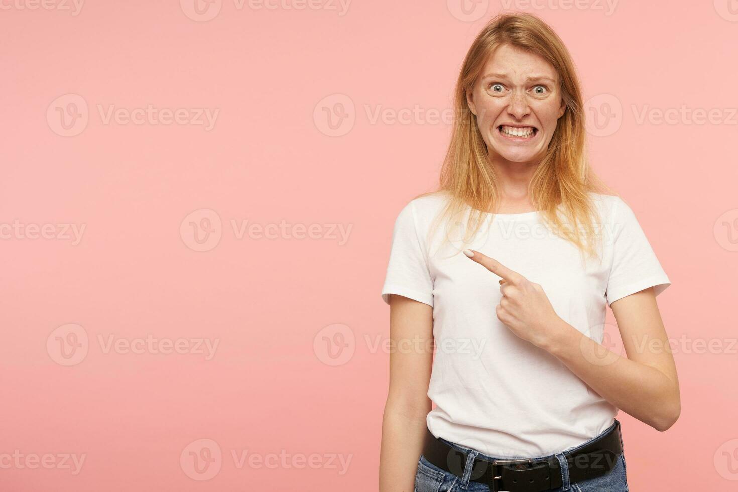Confused young green-eyed foxy lady with casual hairstyle grimacing her face while pointing aside with raised forefinger, isolated against pink background photo