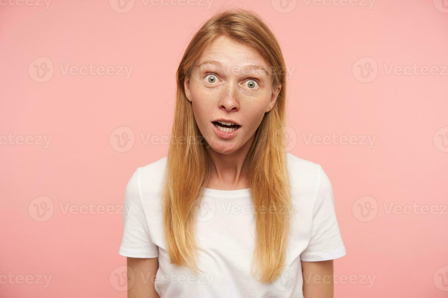 Surprised young lovely long haired redhead female with natural makeup looking wonderingly at camera with wide eyes opened, standing over pink background photo