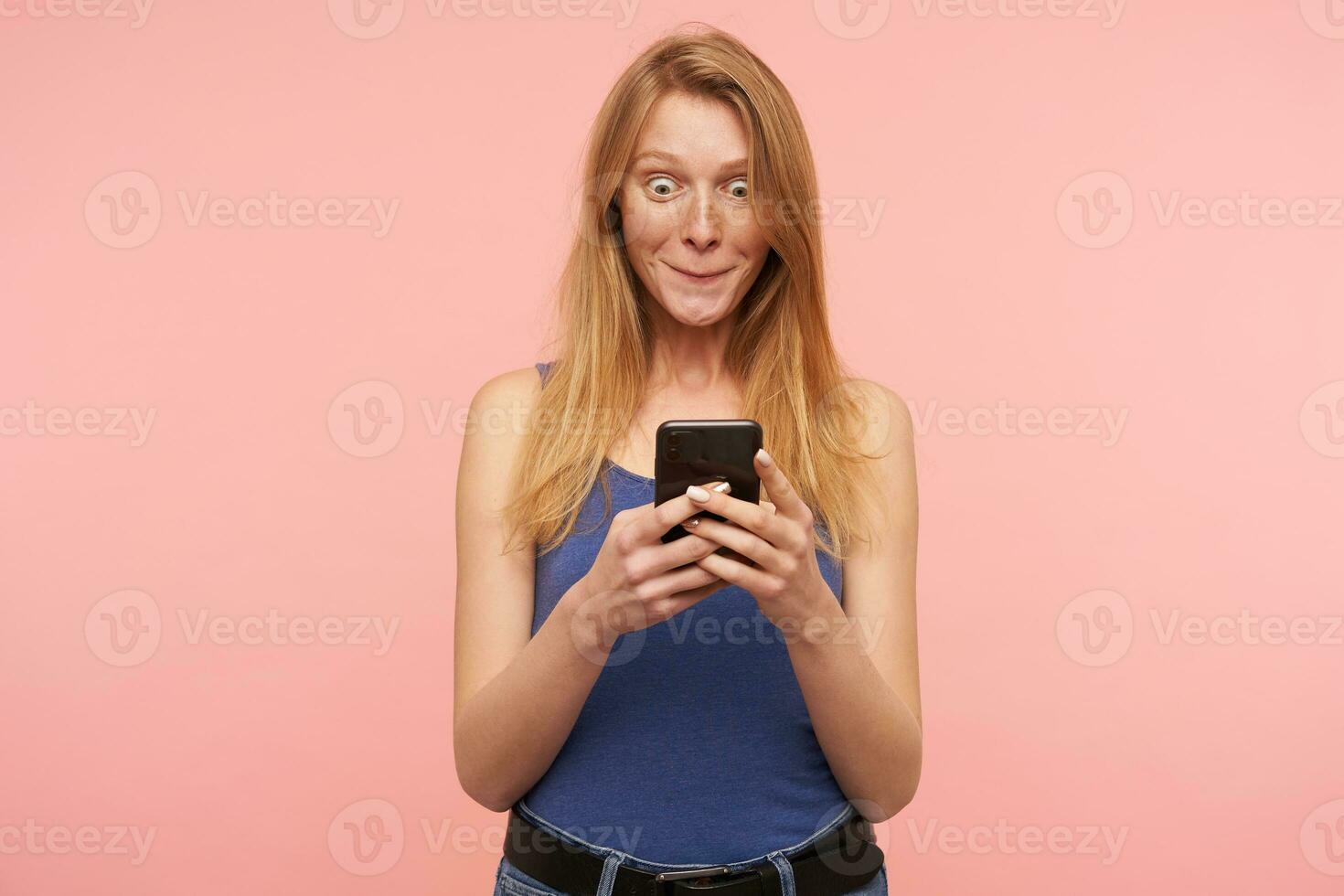 Indoor photo of young lovely redhead woman with casual hairstyle keeping mobile phone in raised hands and being surprised while reading message, isolated over pink background