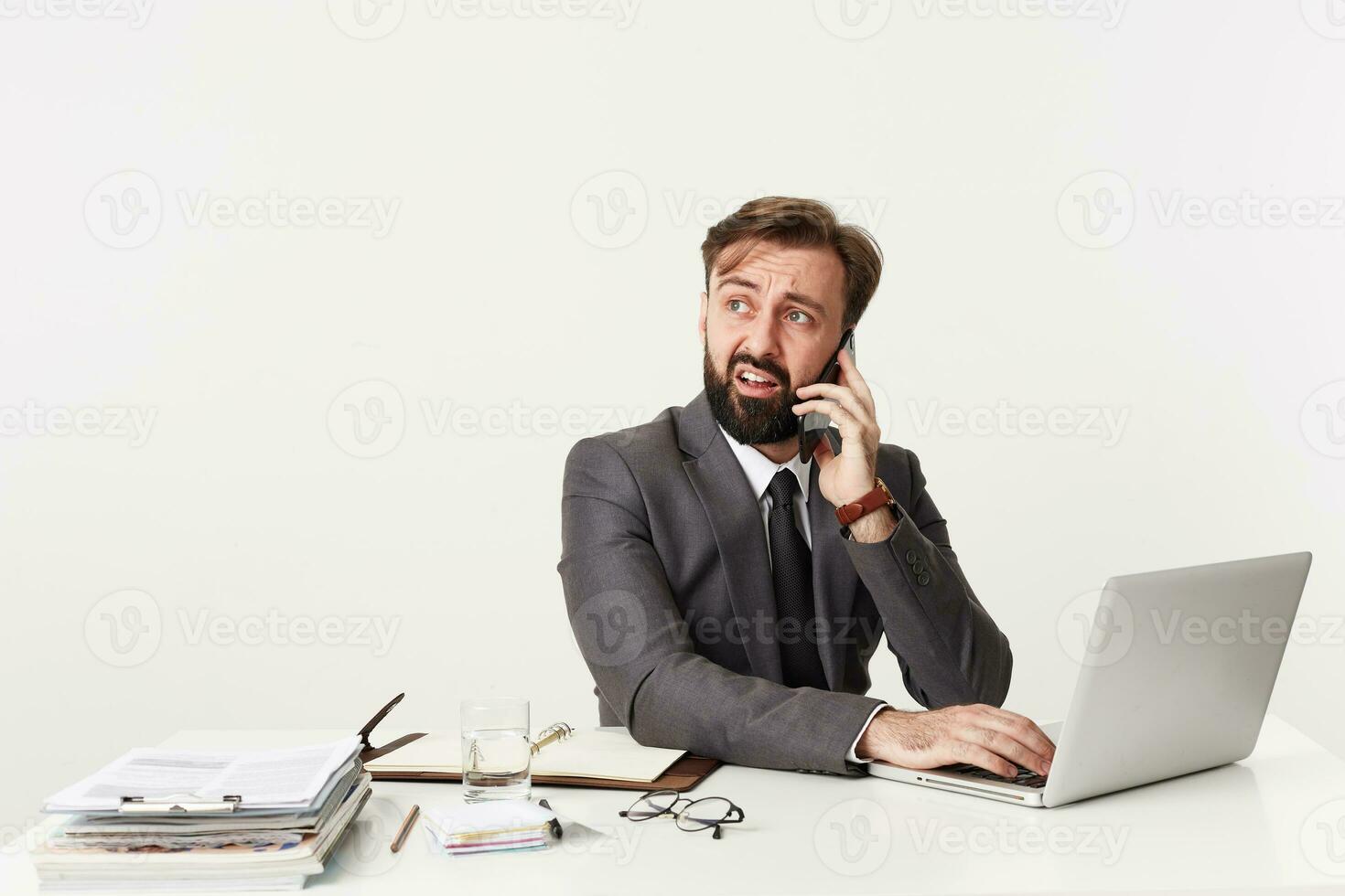 Puzzled young brunette man with beard looking aside with confused face and wrinkling his forehead, making call with smartphone while sitting at table with hands on keyboard of his laptop photo