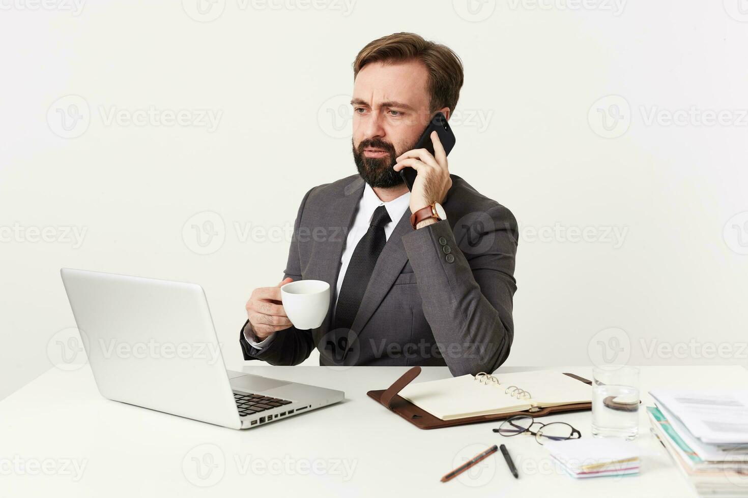 Serious bearded brunette man in formal clothes keeping white cup in raised hand while having phone conversation, frowning eyebrows while looking at screen of his laptop, isolated over white background photo