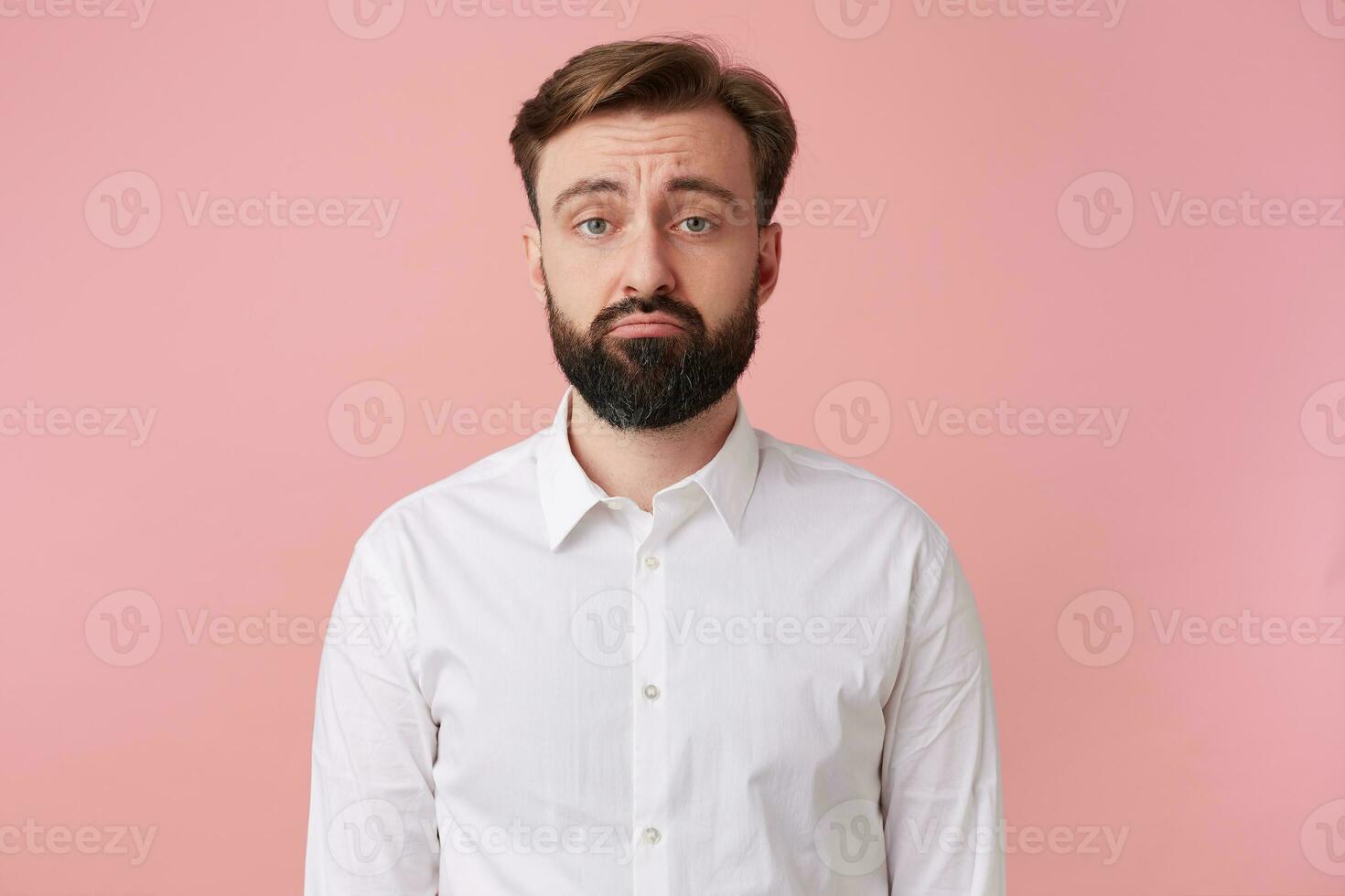 Portrait of a handsome bearded man who is upset about something, wearing a white shirt. Looking at camera with sullen facial expression isolated over oink background. photo