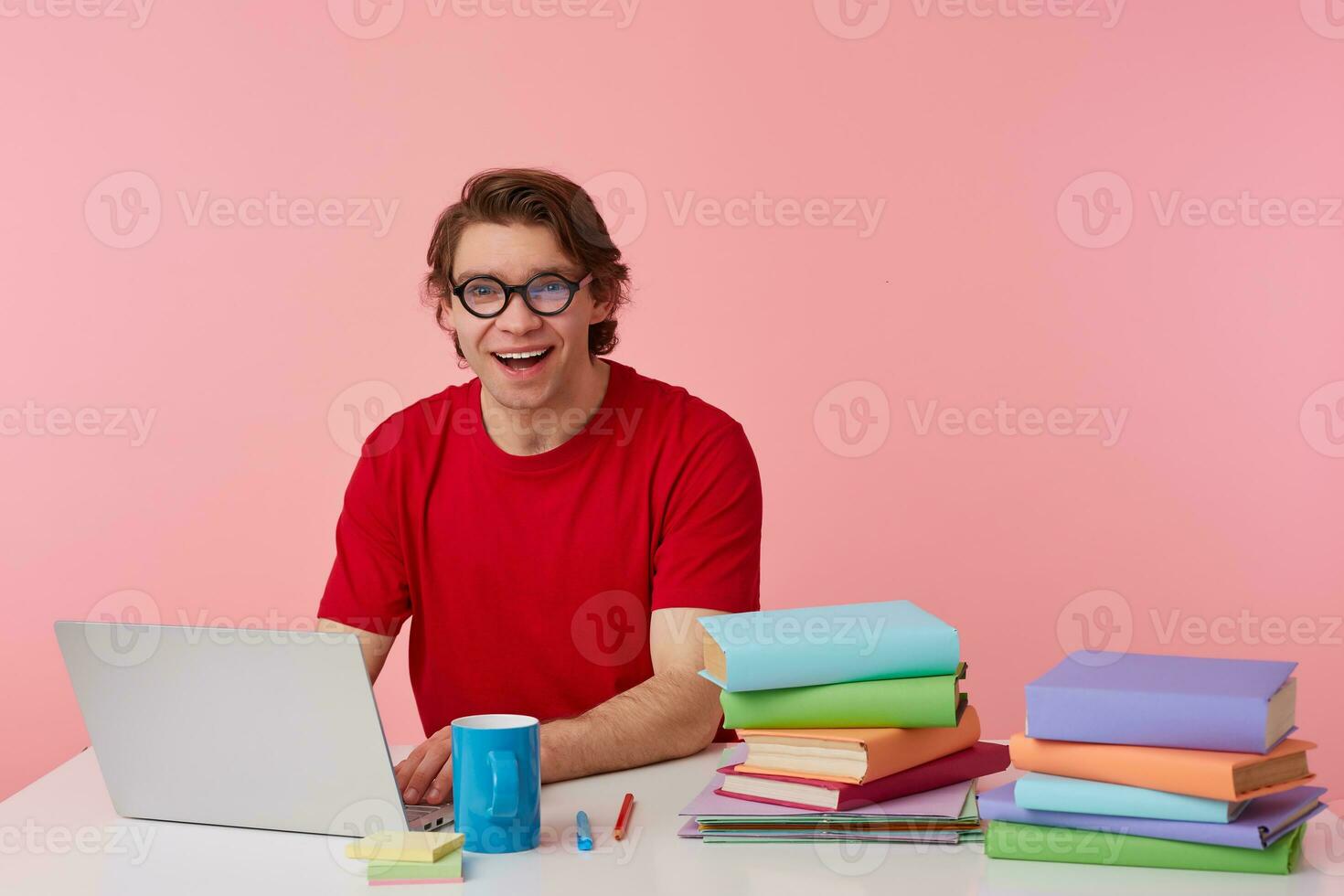 Photo of happy young student in glasses wears in red t-shirt, man sits by the table and working with laptop and books, broadly smiles, looks glad, isolated over pink background.