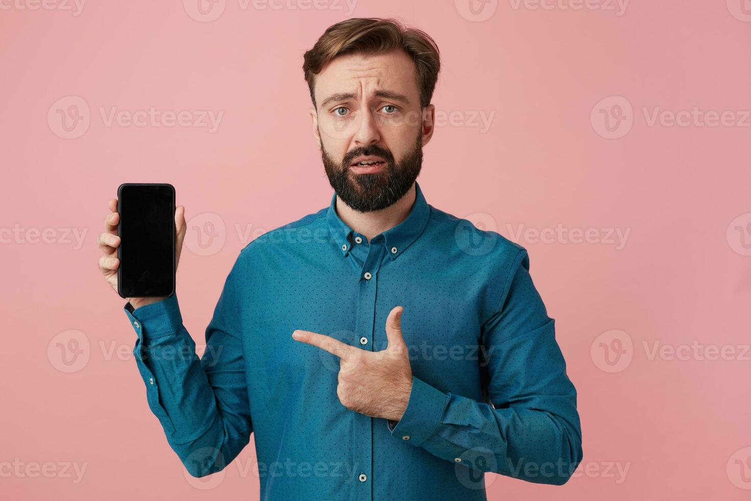 Frowning unhappy attractive bearded man, looking at camera, upset that his smartphone is outdated, wearing a denim shirt, pointing with finger to his device. photo