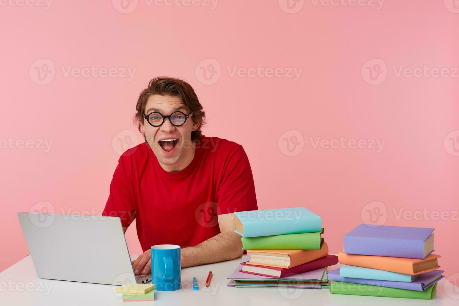 Portrait of happy amazed young student in glasses wears in red t-shirt, man sits by the table and working with laptop and books, looks surprised, isolated over pink background. photo