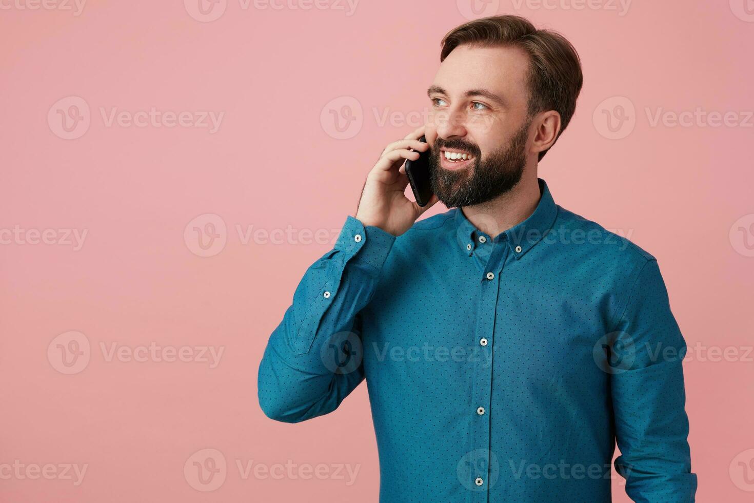 Glad attractive bearded man, looking away and smiling, wearing a denim shirt, talking on the phone. Isolated over pink background. photo