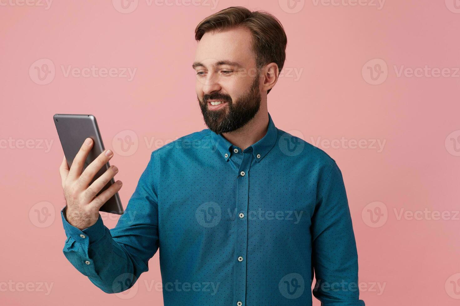 Young attractive bearded man dressed in a denim shirt, smiling, talking to his mom on video chat. Isolated over pink background. photo