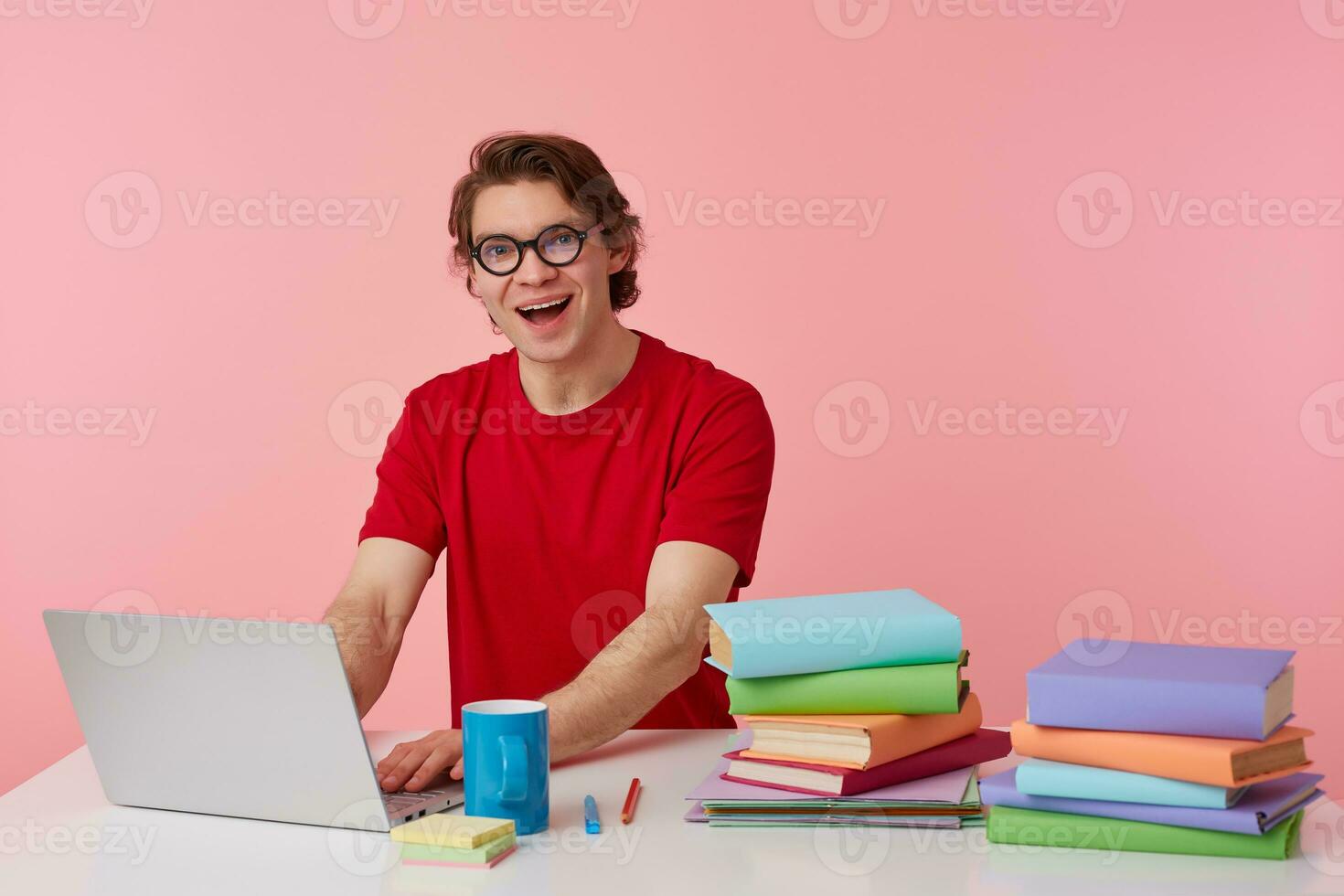 Young positive man student in glasses wears in red t-shirt, sits by the table and working with books and notebook, prepared for exam, having serious look, broadly smiles isolated over pink background. photo