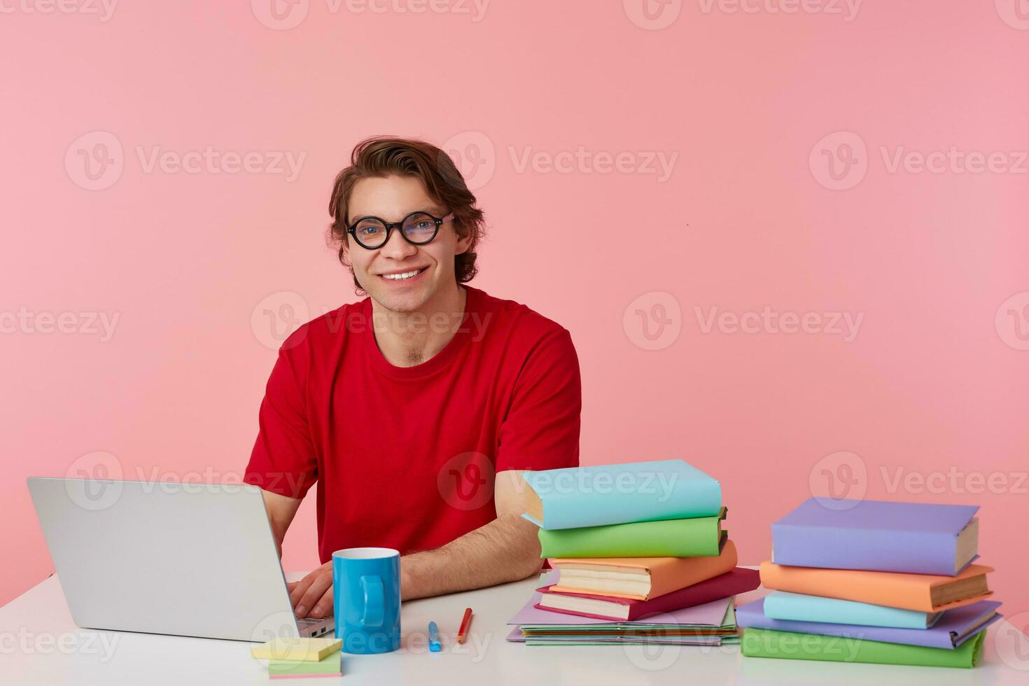 Portrait of smiling young student in glasses wears in red t-shirt, man sits by the table and working with laptop and books, broadly smiles, looks cheerful, isolated over pink background. photo
