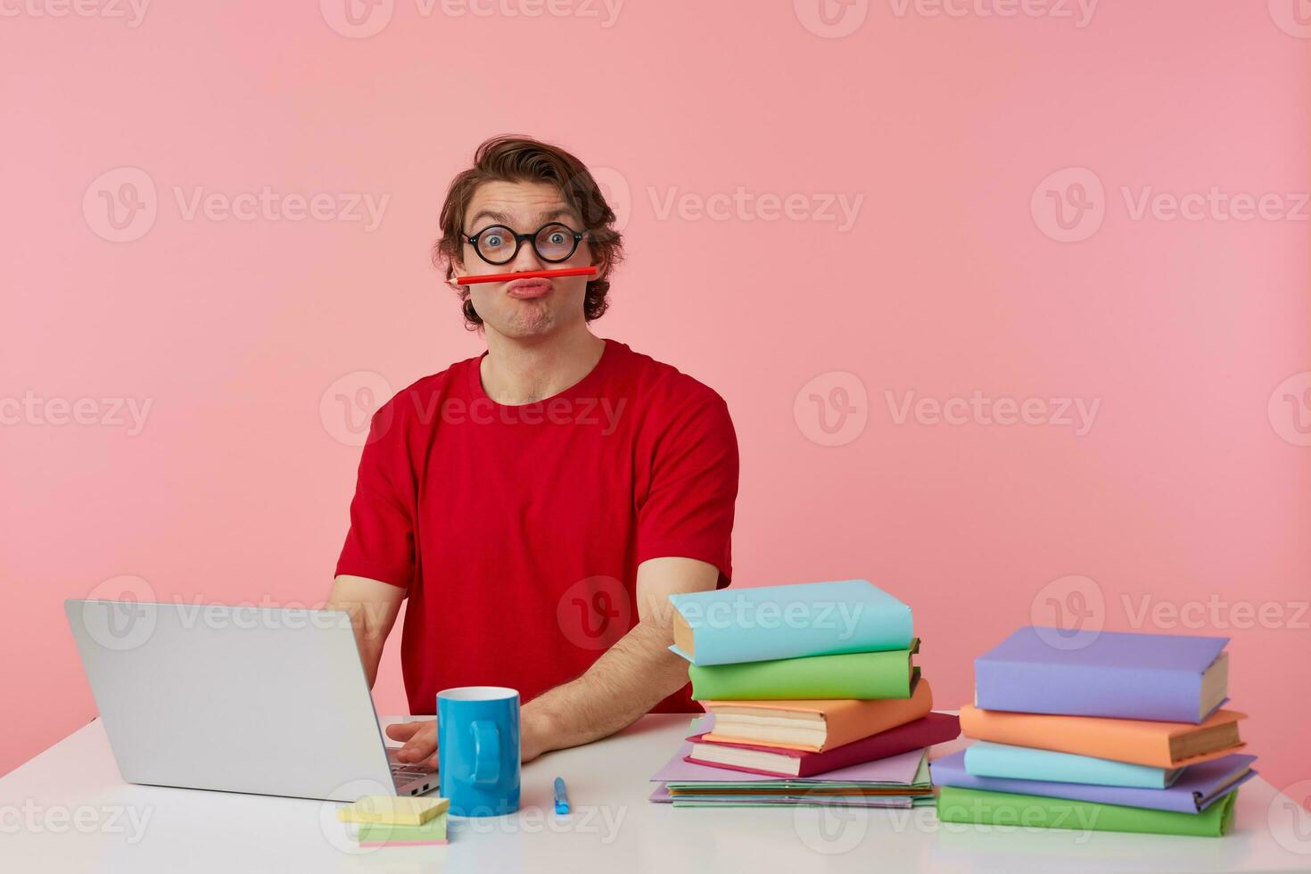Portrait of funny young student in glasses wears in red t-shirt, sits by the table and working with laptop, looks happy and joyful, holding a pencil with his lips isolated over pink background. photo