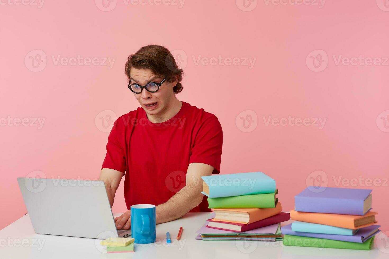Portrait of shocked young student in glasses wears in red t-shirt, sits by the table and working with laptop, looks at the monitor with disgusted, looks unhappy, isolated over pink background. photo