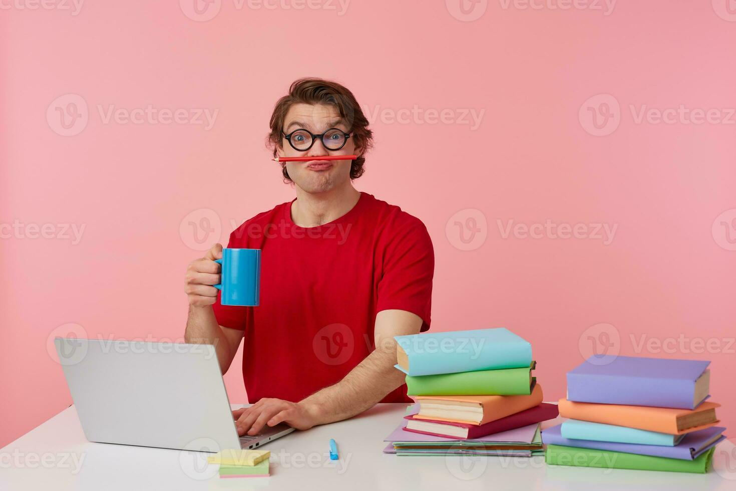 Funny young student in glasses wears in red t-shirt, sits by the table and working with laptop, looks happy and joyful, holding a pencil with his lips and cup in hand, isolated over pink background. photo