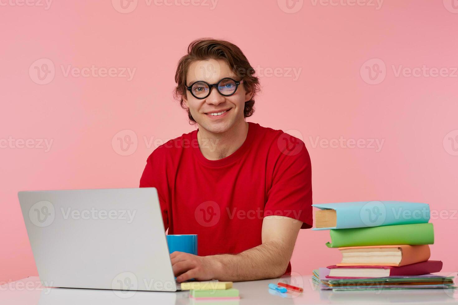 Photo of cheerful young student man in glasses wears in red t-shirt, sits by the table and working with laptop and books, broadly smiles, looks glad, isolated over pink background.