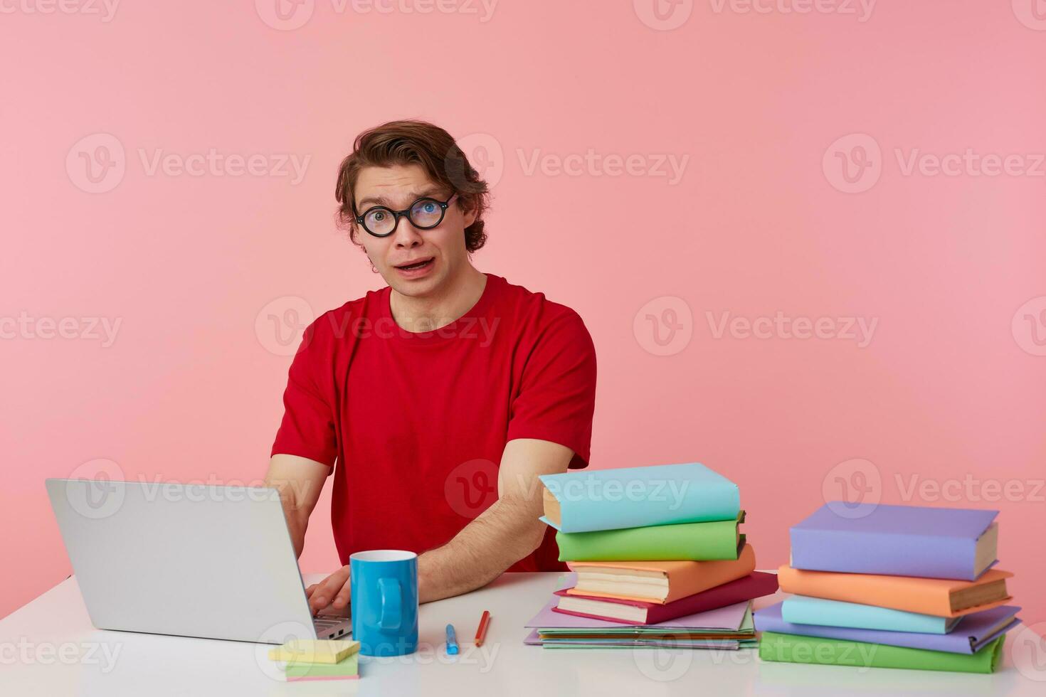 Portrait of sad young student in glasses wears in red t-shirt, sits by the table and working with laptop, looks unhappy, isolated over pink background. photo