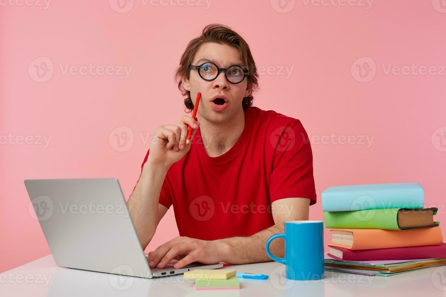 Thinking young man in glasses wears in red t-shirt, sits by the table and working with laptop, looks up, holds in hand a pencil, trying to solve a hard equation, isolated over pink background. photo