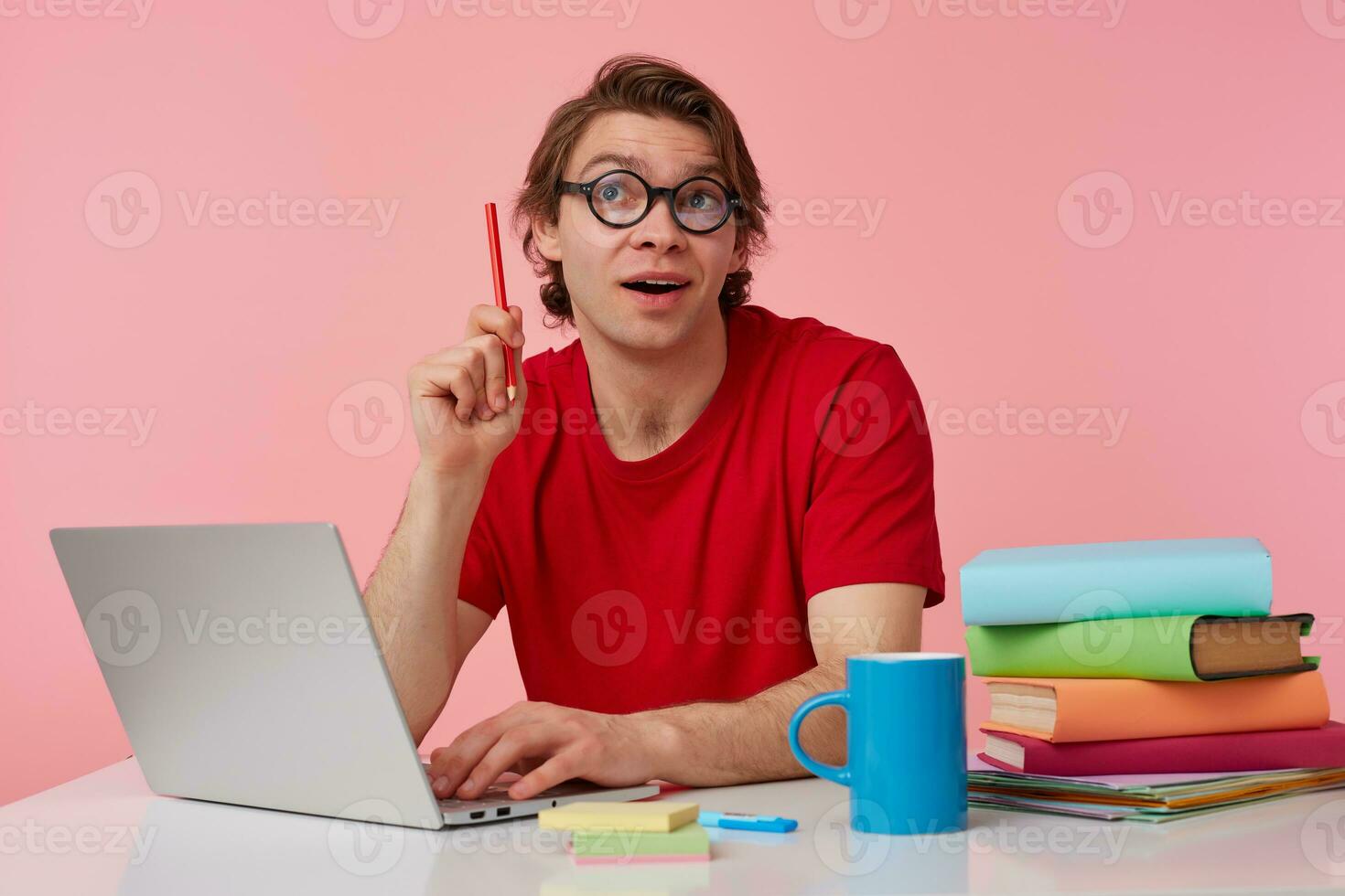 Thinking man in glasses wears in red t-shirt, sits by the table and working with laptop, looks up, holds in hand a pencil, have a idea how to solve a hard equation, isolated over pink background. photo