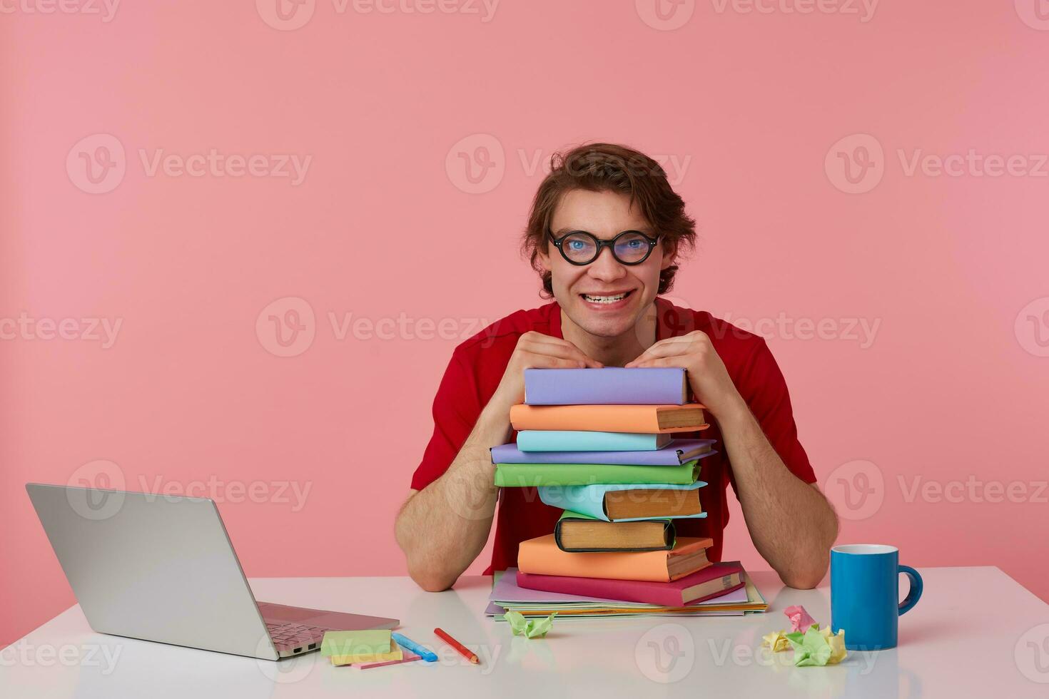 Crazy young man in glasses, in red t-shirt, man sits by the table and working with laptop and books, leaned on a pile of books, looks evil. Isolated over pink background. photo