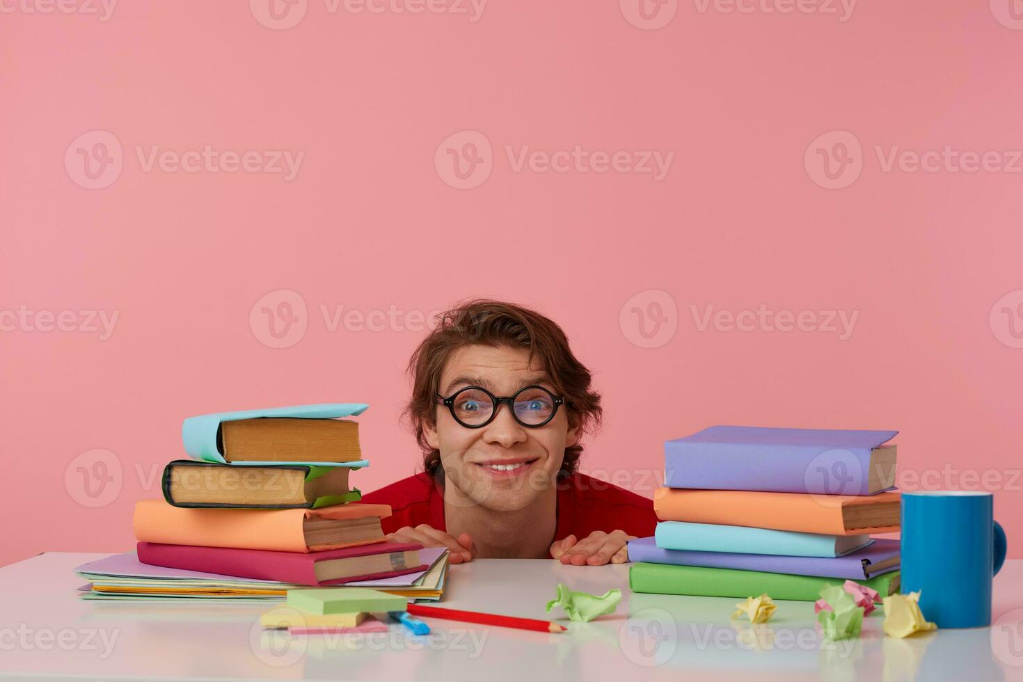Positive man in glasses wears in red t-shirt,hiding at the table with books, looks at the camera and smiling, isolated over pink background. photo