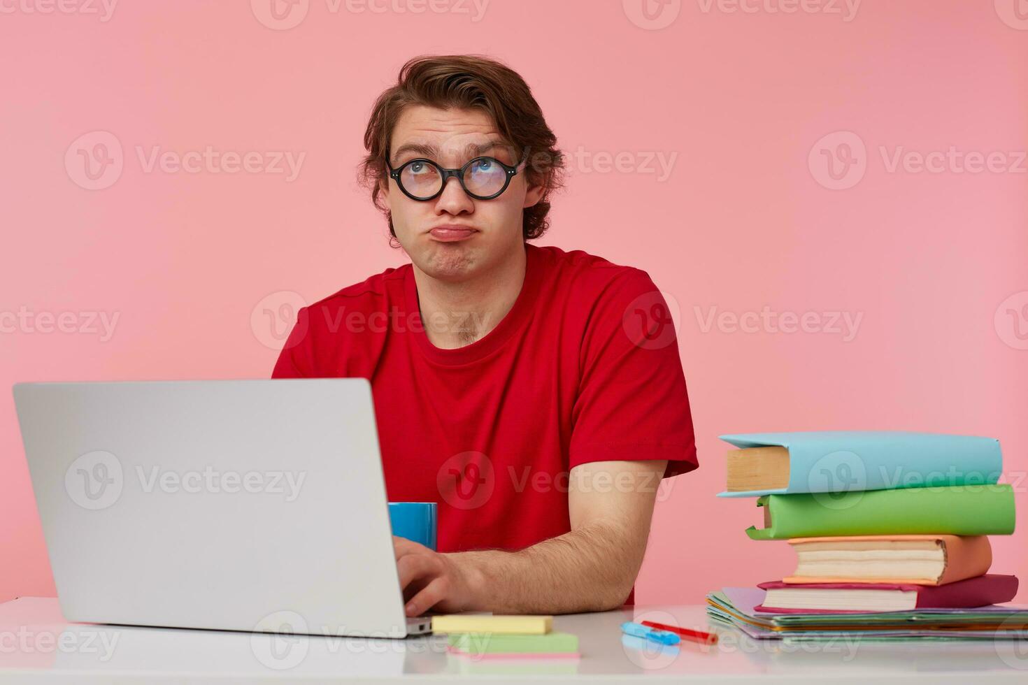 Thinking young student in glasses wears in red t-shirt, man sits by the table and working with laptop, looks up and doubt, isolated over pink background. photo
