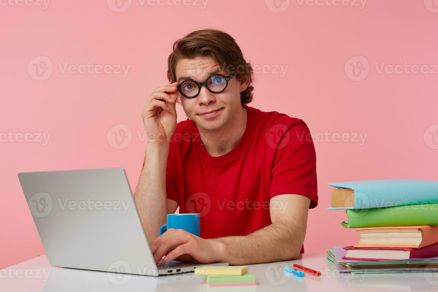 Young student in glasses wears in red t-shirt, man sits by the table and working with laptop, looks at the camera through glasses with confused expression, isolated over pink background. photo