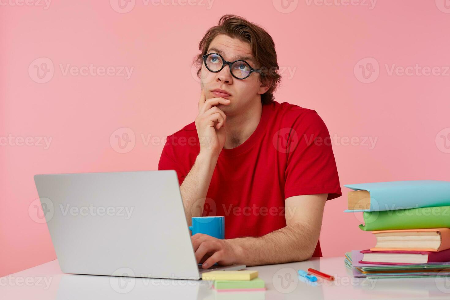 Thinking young student in glasses wears in red t-shirt, man sits by the table and working with laptop, touches chin, looks up and dreaming about holidays, isolated over pink background. photo