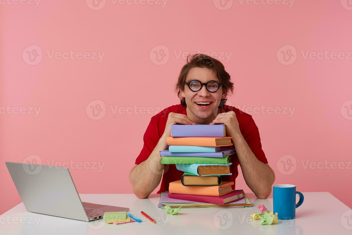Smiling young man in glasses wears in red t-shirt, man sits by the table and working with laptop and books, leaned on a pile of books, looks happy and glad. Isolated over pink background. photo