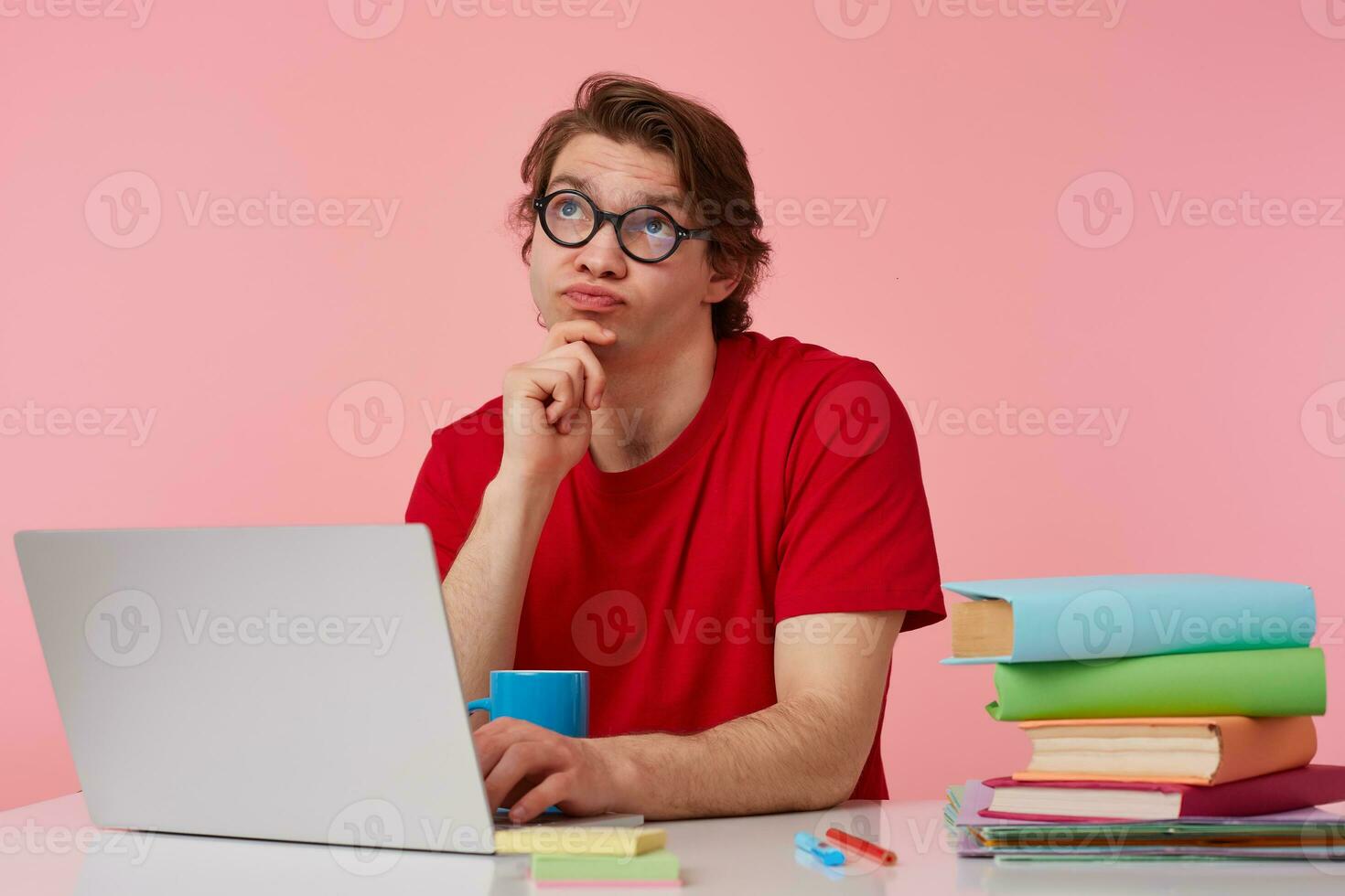 Thinking young student in glasses wears in red t-shirt, man sits by the table and working with laptop, looks up and dreaming about holidays, isolated over pink background. photo