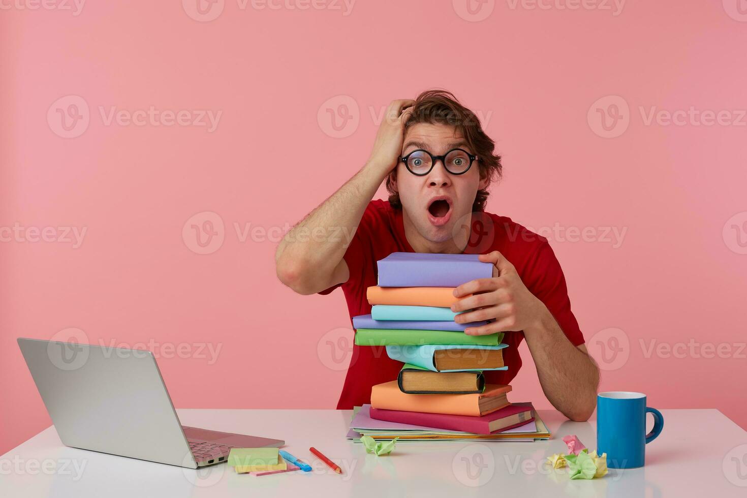 Photo of young shocked guy in glasses sits by the table and working with laptop and books, leaned on a pile of books, looks surprised with wide open mouth. Isolated over pink background.