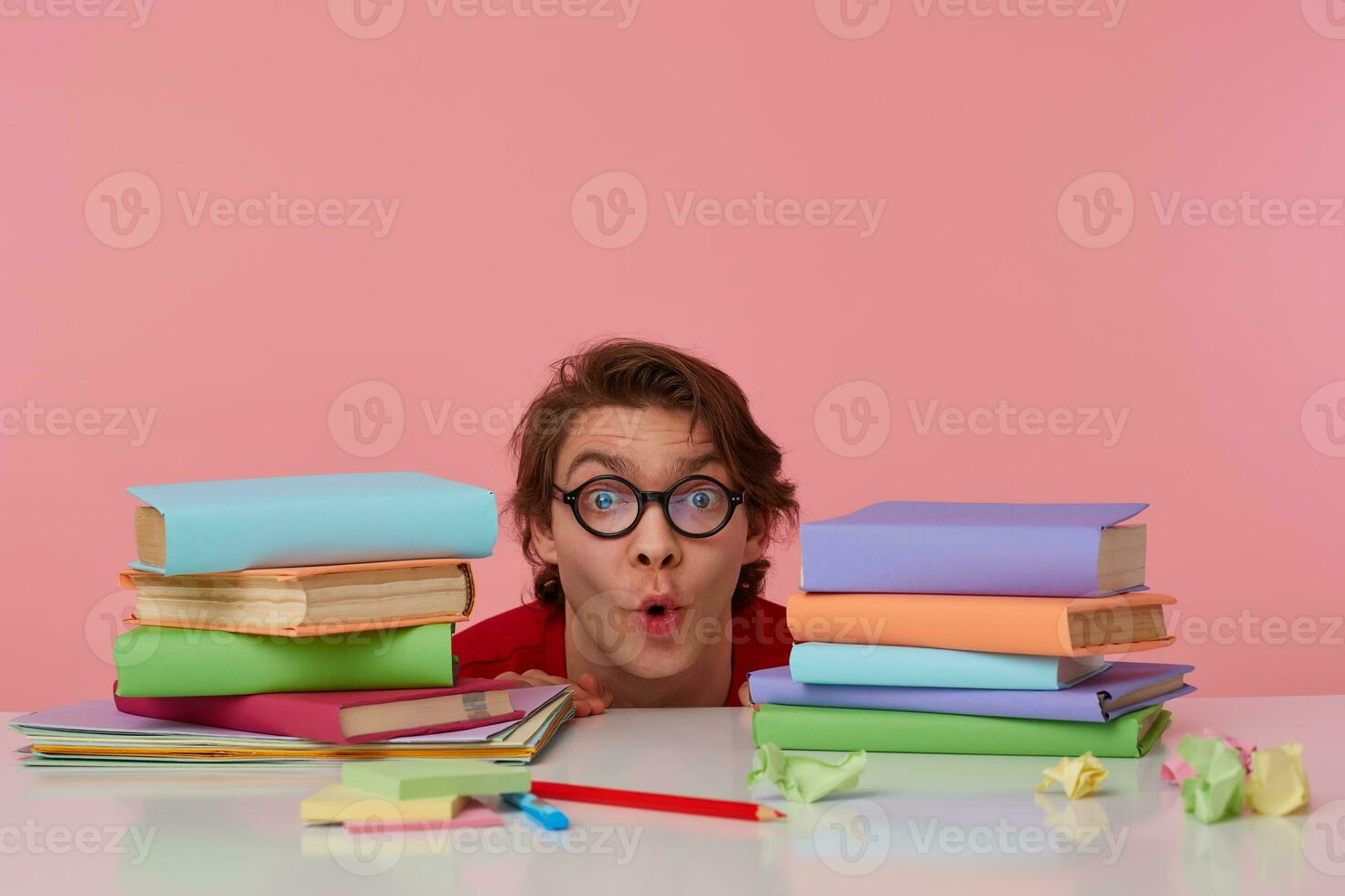 Portrait of happy amazed man in glasses wears in red t-shirt, hiding at the table with books, looks at the camera with surprised expression, looks cheerful, isolated over pink background. photo