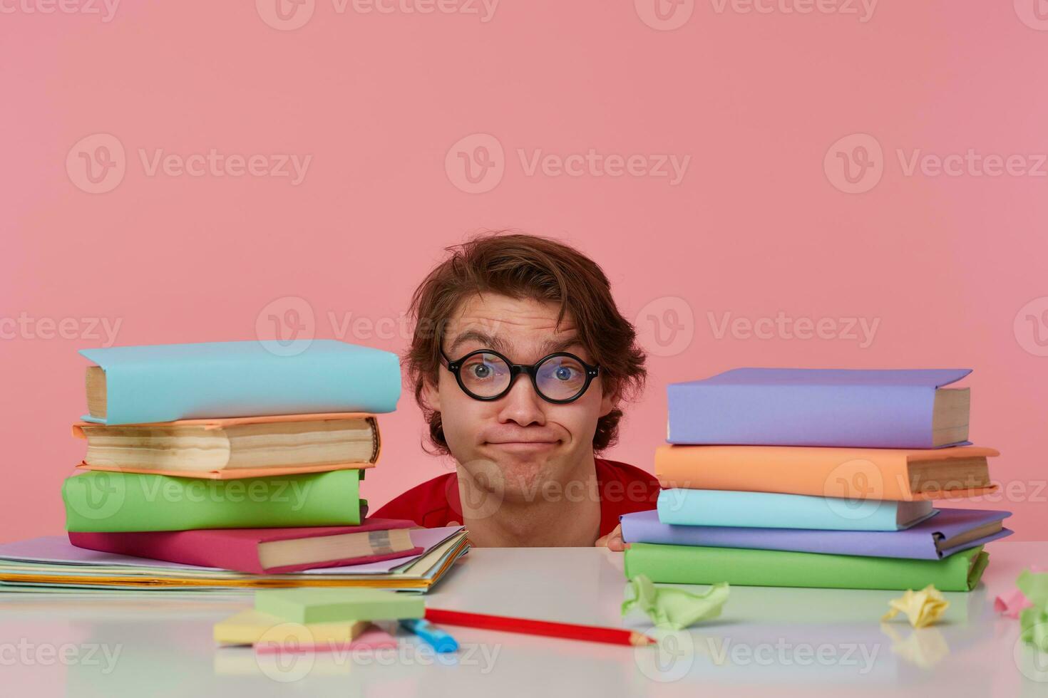 Unhappy young man in glasses wears in red t-shirt, hiding at the table with books, isolated over pink background. Looks displeased and sad. photo