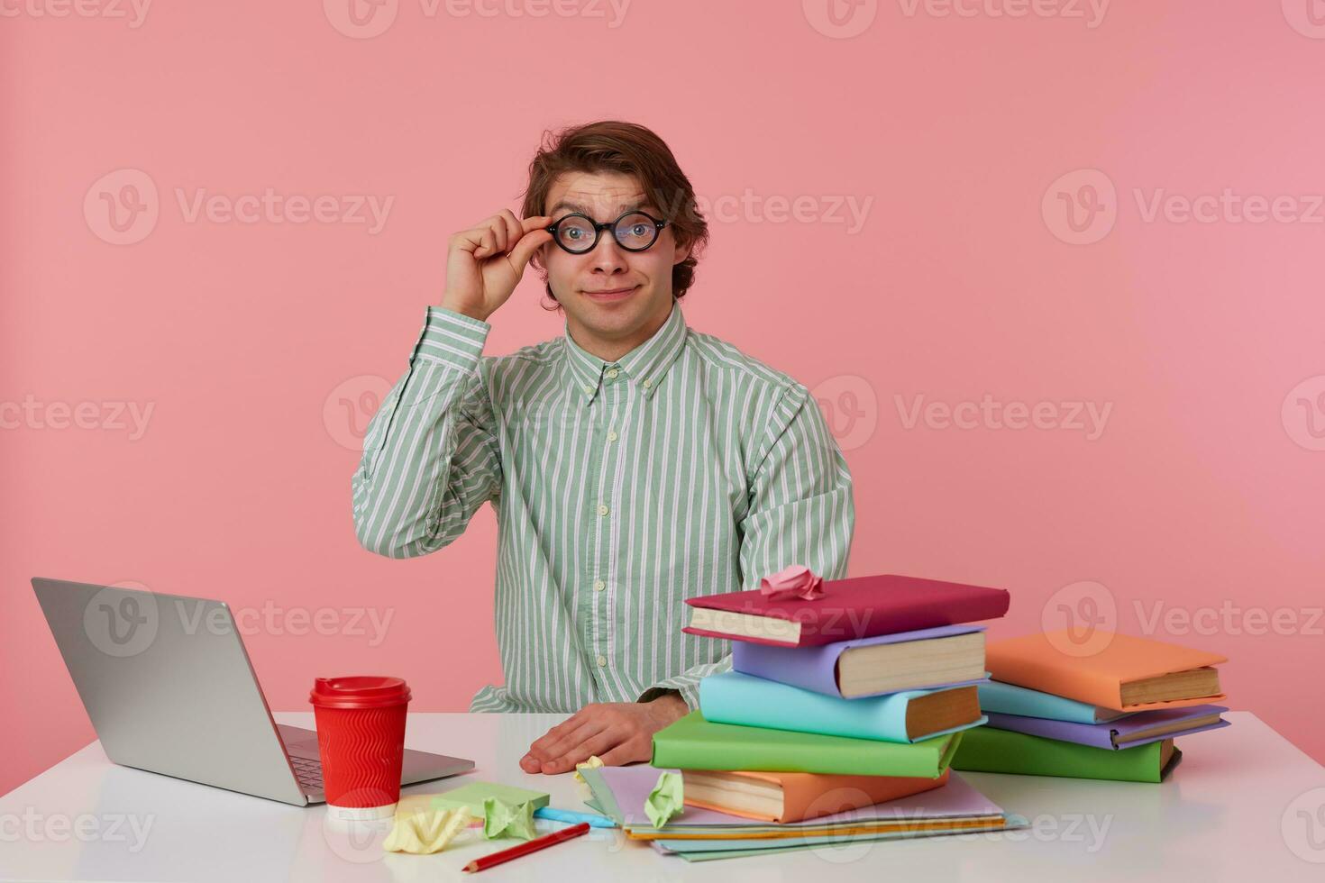 Portrait of smiling man student in glasses wears in red t-shirt, sits by the table and working with notebook and books, prepared for exam, looks through glasses, isolated over pink background. photo