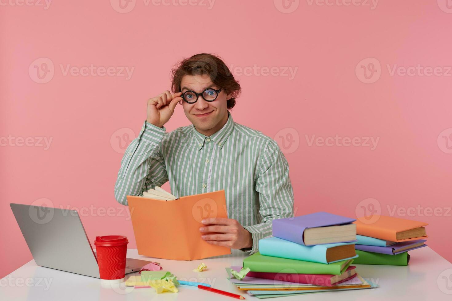 Young cheerful man in glasses wears in shirt, sits by the table and reads book, looks at the camera through glasses, working with notebook, prepared for exam, isolated over pink background. photo