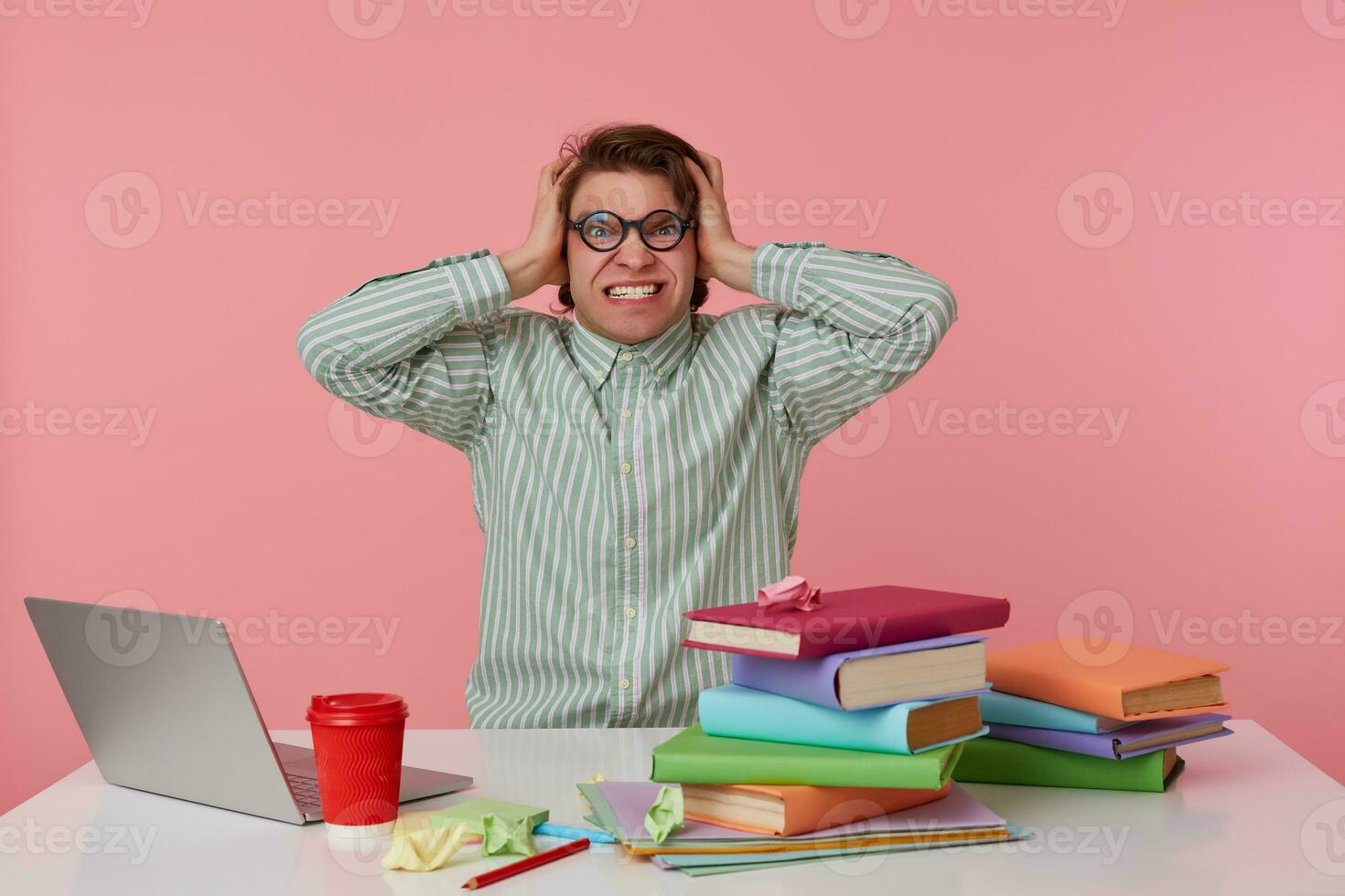 Young angry student in glasses, sits by the table and working with laptop, holds his head and looks surprised , isolated over pink background. photo