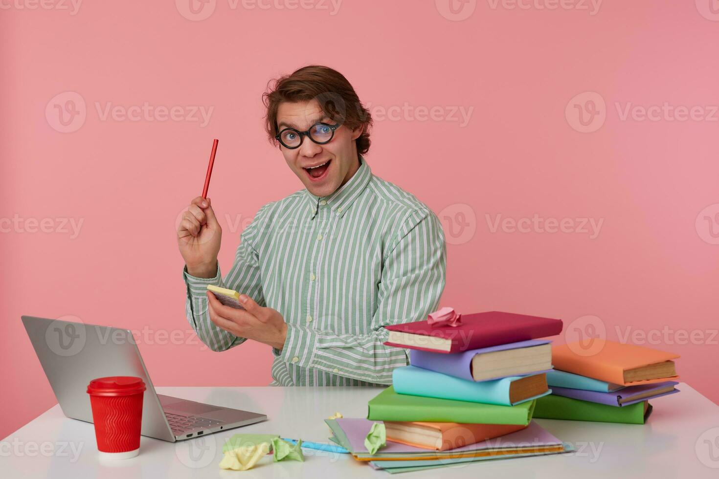 Young guy in glasses sits by the table and working with laptop, looks at the camera, holds in hand a pencil and stickers, have a great idea, isolated over pink background. photo