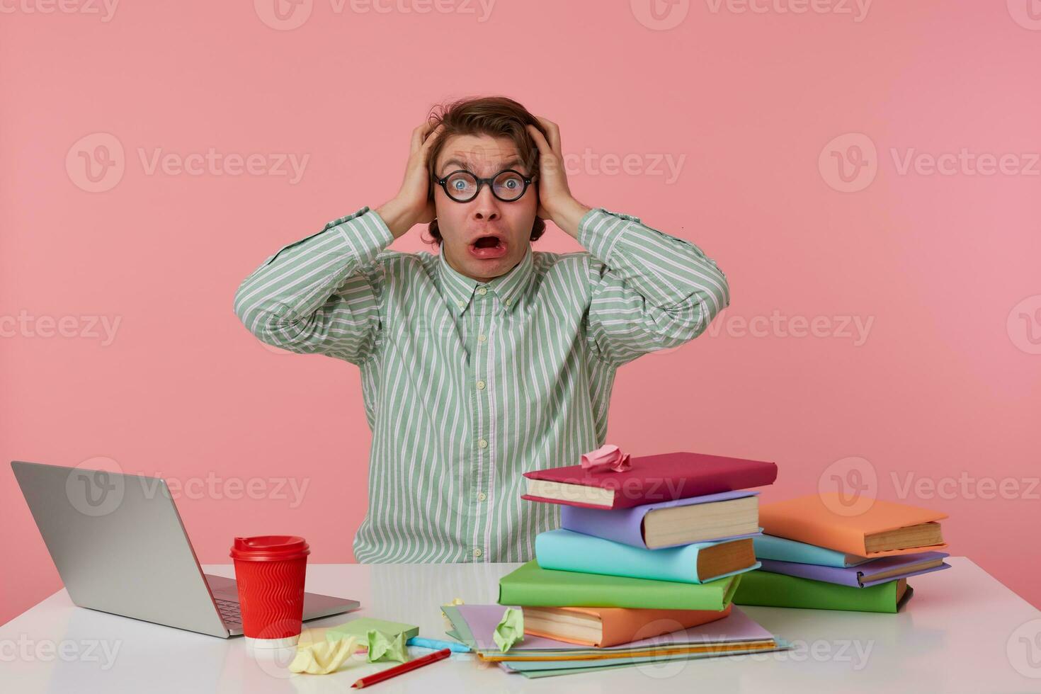Portrait of young angry man in glasses, sits by the table and working with laptop, holds his hesd and looks surprised , isolated over pink background. photo