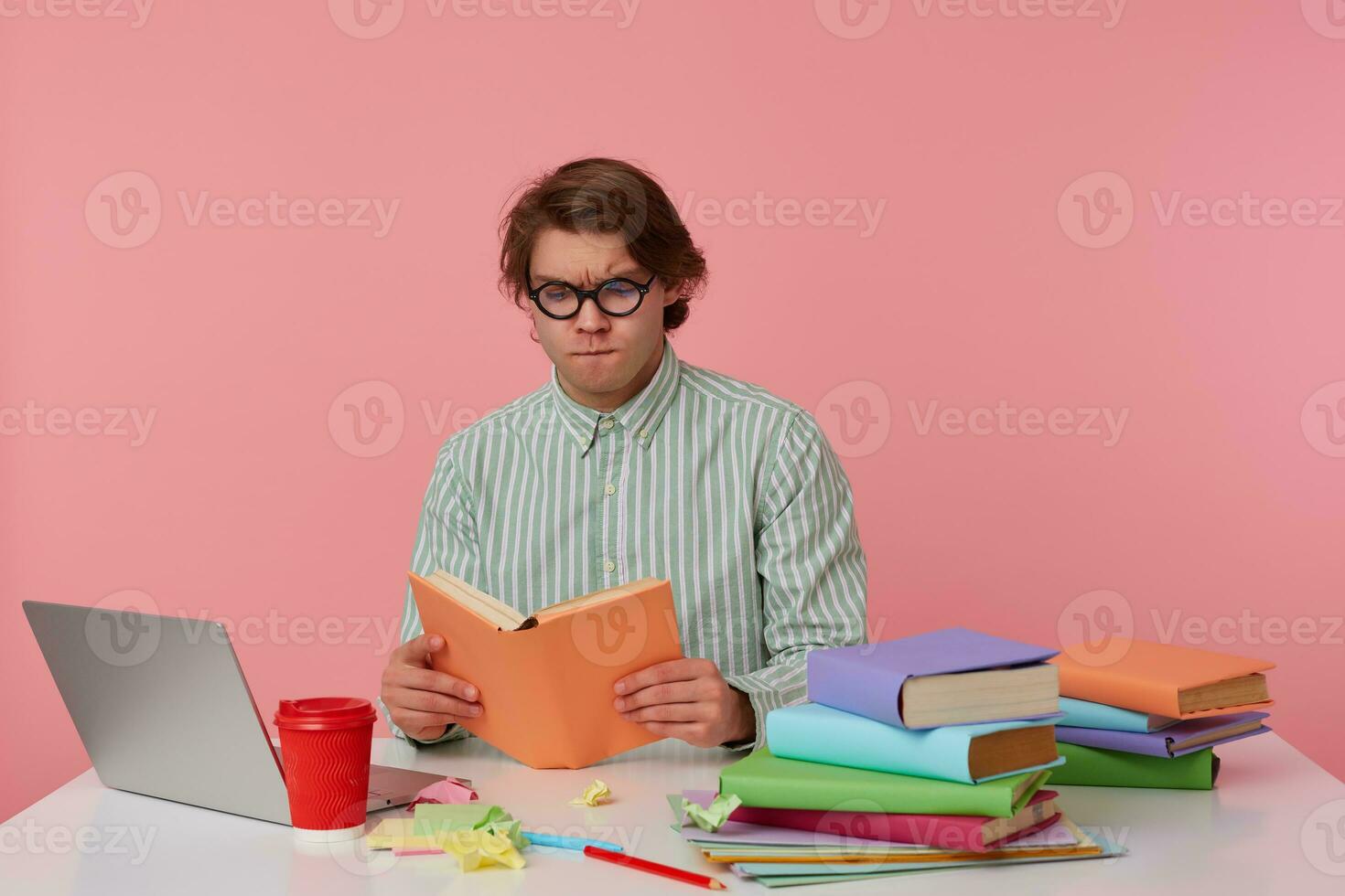 Young concentrated man in glasses wears in shirt, sits by the table and working with notebook, prepared for exam, reads book, looks serious, isolated over pink background. photo