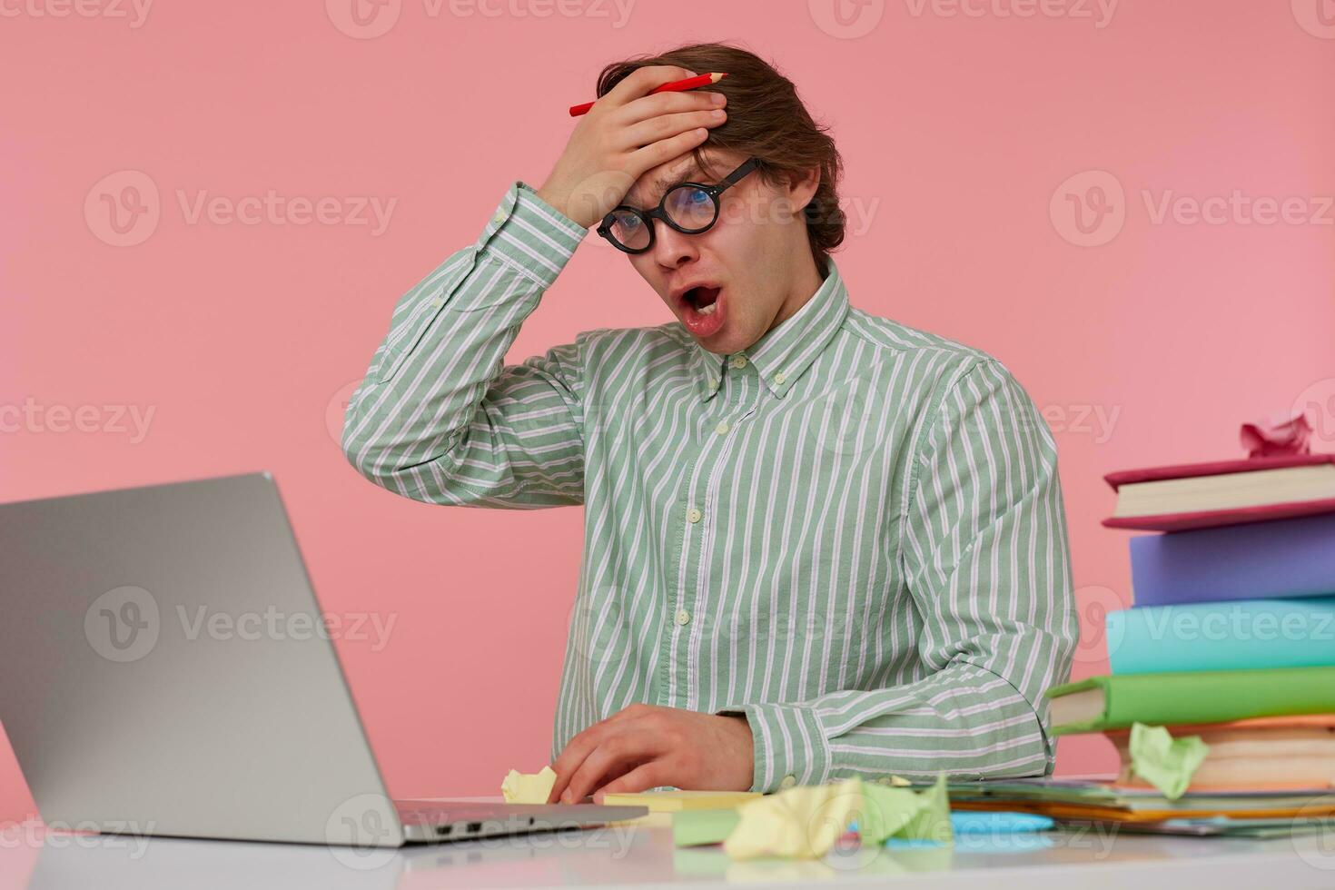 Portrait of man in glasses sits by the table and working with laptop, very dissatisfied, holds hand on forehead with wide open mouth, isolated over pink background. photo