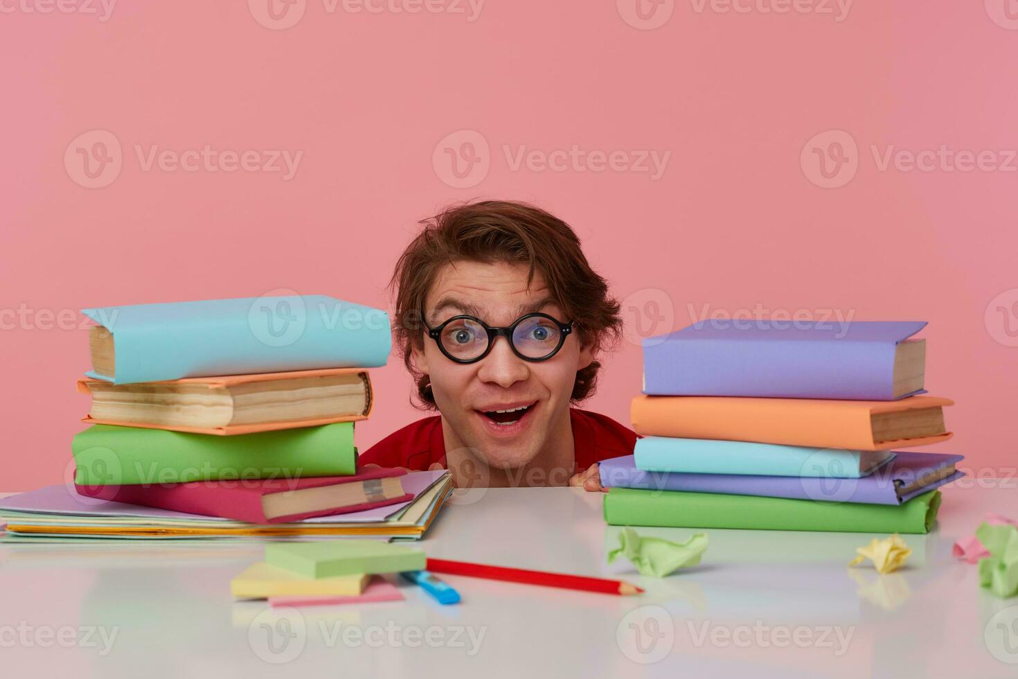 Photo of happy amazed guy in glasses wears in red t-shirt, hiding at the table with books, looks at the camera with wide open mouth, looks cheerful, isolated over pink background.