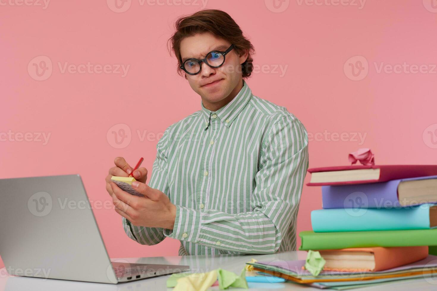 Young doubtful man in glasses sits by the table and working with laptop, holds in hand a pencil, frowning and writes the idea on the sticker, isolated over pink background. photo